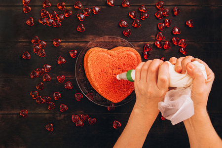 Women's hands squeeze cream from a cooking bag onto a red heart-shaped cake. Preparation for Valentine's Day.の写真素材