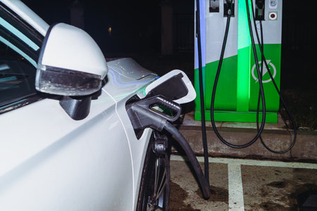The white car is connected to the electric charging station at night in neon lights in the rain.