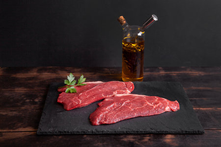 Raw minute steak of marbled beef with a parsley leaf on a stone slate board, glass bottle with vegetable oil and balsamic vinegar.の写真素材