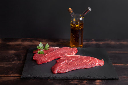Raw minute steak of marbled beef with a parsley leaf on a stone slate board, glass bottle with vegetable oil and balsamic vinegar.の写真素材