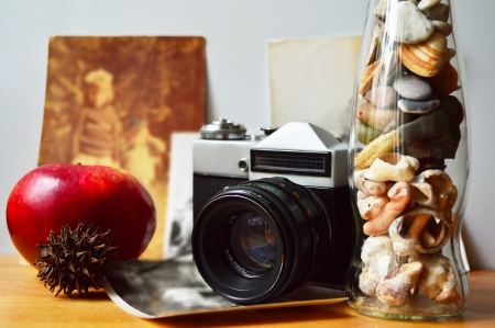 A stilllife with old camera, old photos, a fresh apple and a glass bottle with shells and stones