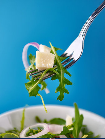 A close-up of a fork with a vibrant salad, contrasted with a striking blue background, emphasizing the freshness. Parmesan shavings, shallots and arugula.の写真素材