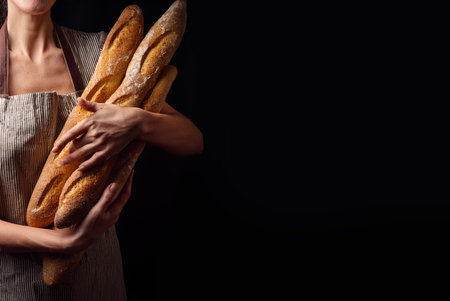 A young woman dressed in a baker's apron holds freshly baked crusty baguettes with subtle care. Text space.の写真素材