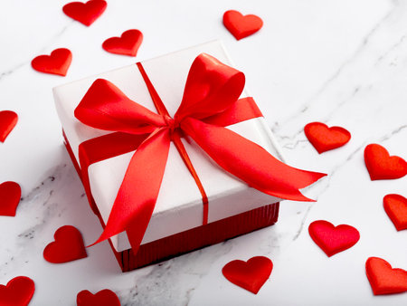 Gift box with big vibrant red ribbon and scattered small red heart decorations on a white marble background. Perfect for Valentine's Day or romantic occasion themes.の写真素材