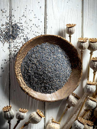 Rustic wooden bowl filled with fresh poppy seeds on a white wooden background, surrounded by dried poppy pods. Perfect for culinary uses and health benefits.の写真素材