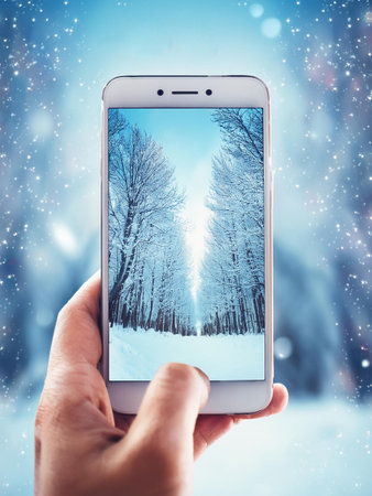 A hand holding a smartphone capturing a winter landscape with snow-covered trees and a bright, clear sky in the background.の写真素材