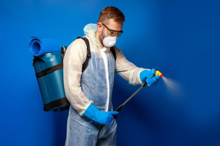 A young man wearing protective gear, including goggles and a mask, uses a spray gun with a backpack tank against a blue background.の写真素材