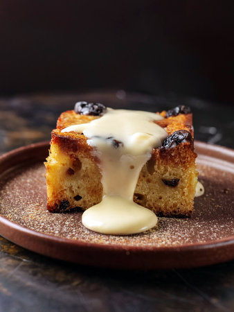 A slice of bread pudding topped with creamy vanilla sauce and raisins on a brown plate, with a bowl of extra sauce in the background. Perfect dessert for a cozy homemade treat.の写真素材