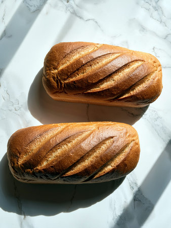 Two freshly baked loaves of bread with golden crusts on a marble countertop, highlighted by sunlit stripes, showcasing artisanal baking.の写真素材