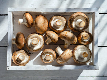 Wooden crate filled with fresh brown mushrooms placed on a white wooden surface, with natural sunlight creating a pattern of shadows. Perfect image for articles on organic produce,の写真素材