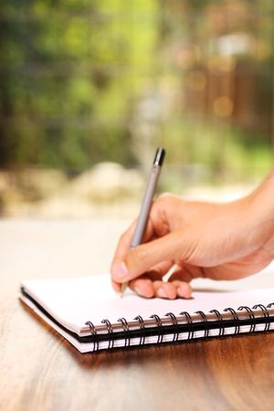 A hand writing on a notepad on a table, in front of a window.