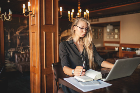 Business woman in glasses indoor with coffee and laptop taking notes in restaurantの写真素材