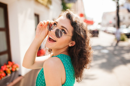Young woman in round sunglasses and dress with curly hair smiling over the shoulder in the city streetの写真素材