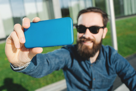 Young hipster in sunglasses and beard making selfie with coffee outdoors in the cityの写真素材