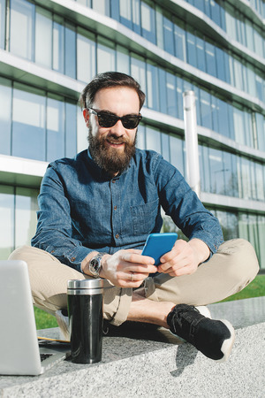 Young hipster in sunglasses and beard making selfie with coffee outdoors in the cityの写真素材