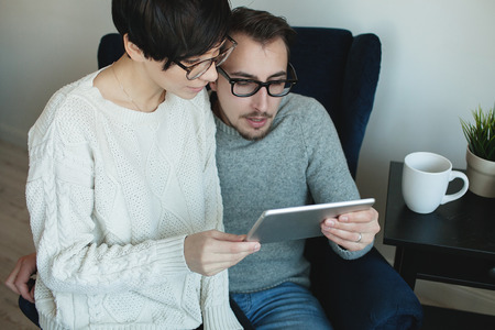 Young hipster couple in eyewear enjoying the tablet together sitting on the chair in apartmentの写真素材