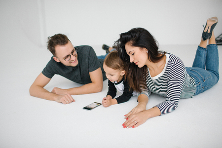 Young hipster father, mother holding cute baby boy on concrete floor with smartphone over white backgroundの写真素材