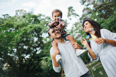 Happy young family playing with bubble wands with son sitting on fathers shoulders in park outdoors in summerの写真素材