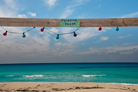 Sign reading Paradise Found on a piece of wood with colorful lights hanging about a beautiful beach in the caribbeaの写真素材