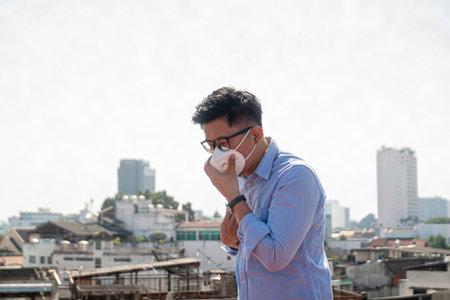 Portrait of young Asian businessman wearing face mask and coughing outdoors.の写真素材