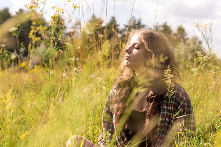 Beautiful young woman with closed eyes and long curly brown hair dressed in a check shirt is sitting on a meadow in high grass and gentle wild flowersの写真素材