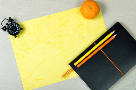 Black notebook, sheet of handmade paper, color pencils, clock and fresh mandarin on light wooden background. Colorful work space. Desk dining.Top view. Flat lay. Tints of yellow and orange.の写真素材