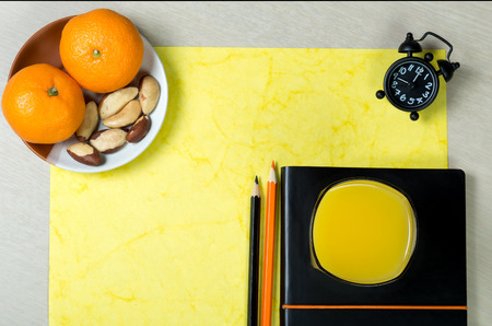 Black notebook, sheet of handmade yellow paper, color pencils, clock, glass of orange juice and healthy snack on light wooden background. Colorful work space. Desk dining.Top view. の写真素材