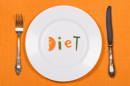 White plate with word diet made of pieces of vegetables on orange background. Vintage fork and knife. Flat lay. Top view. Concept of dieting and healthy eating.の写真素材