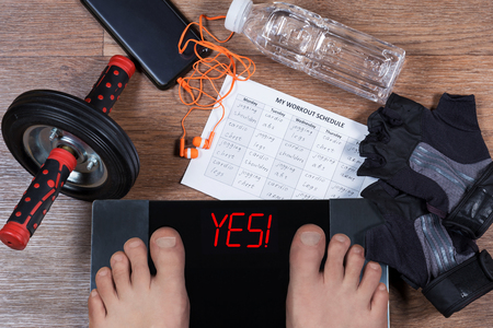 Digital scales with male feet on them and sign "yes!" surrounded by sport accessories (AB roller wheel, workout schedule paper, weightlifting gloves), smartphone with earphones and water bottle. Concept of active and healthy lifestyle.の写真素材