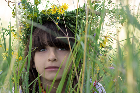 Beautiful little girl with green eyes and a colorful garlang made of wild flowers on her head sits in high green grass on a meadow. Close shot.の写真素材