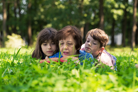 Elderly woman, girl and boy are lying on the lawn and reading a book against green nature background. Grandmother and grandchildren. Concept of harmony relationship in family.の写真素材