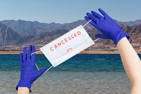 Woman hands with latex gloves on them are holding safety mask with sign cancelled  against beautiful seaside background. Concept of trips cancellation because of coronavirus pandemic.の写真素材