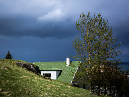 HAFNARFJORDUR, ICELAND - May 31, 2021: 
A scandinavian style house with green roof and chimney, hidden behind green hill. Dark, stormy sky. Hafnarfjordur, Iceland.のeditorial素材