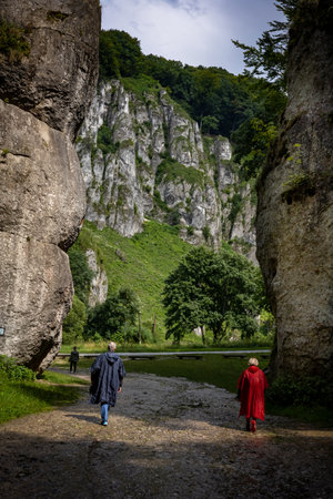 A couple in storm coats walking through a rock gate. 
A picturesque limestone rock formation called "Cracow gateâ. 
Ojcow, Polish Jura, Poland.の写真素材