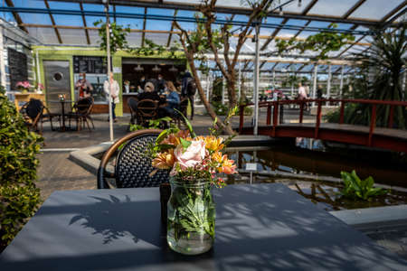 Reykjavik, Iceland - April 26, 2021: Florian Cafe greenhouse bar in Reykjavik's botanical garden. A glass vase with flowers on the table with an empty wicker chair. Sitting people in background.のeditorial素材