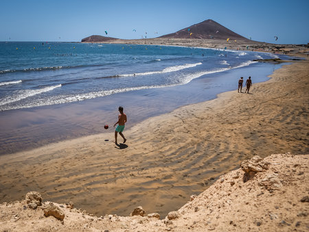 El Medano, Spain - March 19, 2021: A young man playing football on the beach in south Tenerife, Canary Islands. Sunny day, golden sand, Atlantic ocean and mountains in background.のeditorial素材
