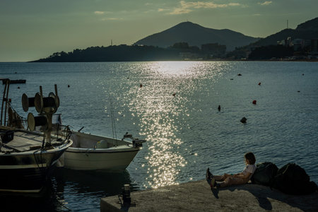 Budva, Montenegro - April 28, 2022: Child sitting on a seaside pier. Small fishing boats floating on water. Scenic mountains in background.のeditorial素材