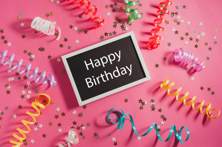Colorful party streamers, gold little stars and blackboard with text Happy Birthday on pink backgrond. の写真素材