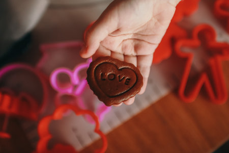 A child's hand holding a heart-shaped cookie dough piece with the word "LOVE" stamped on it. Various red and pink cookie cutters are scattered in the background.の写真素材