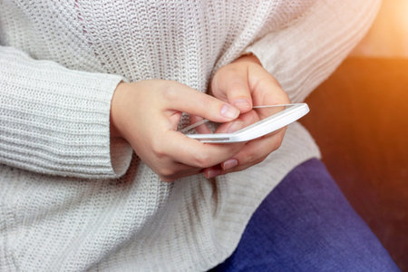 A young woman holding white mobile smartphone in her hands watching video or browsing internet and...