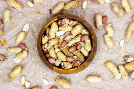 Top view of unpeeled brown peanuts in the round wooden bowl on light background.