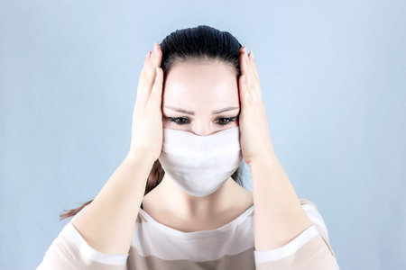 Portrait of sad anxious young caucasian woman wearing white disposable medical face mask on light...