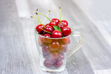 Fresh juicy red sweet cherry berries in the glass cup on light wooden background in summer.