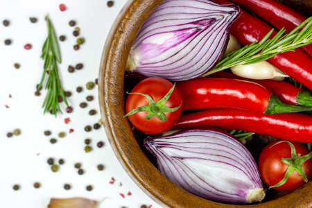 Top view of summer vegetables: onion, garlic, tomato, red chili pepper, rosemary in brown wooden bowl and peppercorns on white table background.の写真素材