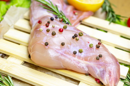 Raw uncooked pink rabbit leg meat with green rosemary, pepper and fresh vegetables on light wooden cutting board background.の写真素材