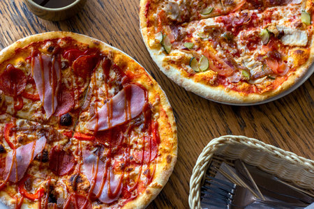 Top view of two fresh baked homemade sliced pizzas with cheese and ham topping served with kitchenware on wooden background.の写真素材