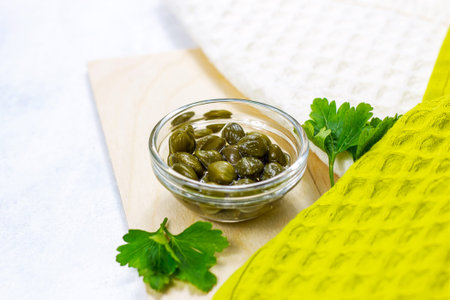 Small green pickled capers fruit in a glass dip bowl on light wooden background in the kitchen.