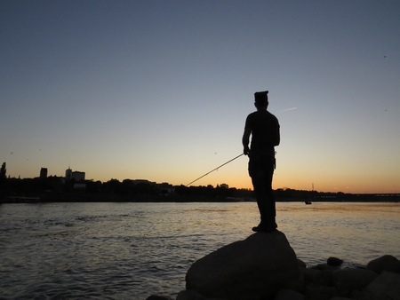 Fisherman Silhouette Standing on Rock with Fishing Rod with a Beautiful City Skyline in Background Seen During Sunsetの写真素材
