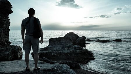 Silhouette of alone man tourist standing on the stone in the sea and enjoying scenery during sunrise or sunset. Cold tones photography.の写真素材