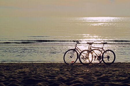 Two bicycles silhouettes standing on the sand near the water edge on the beach. Beautiful shiny and glow water of the sea on the background. Sport and recreation concept.の写真素材